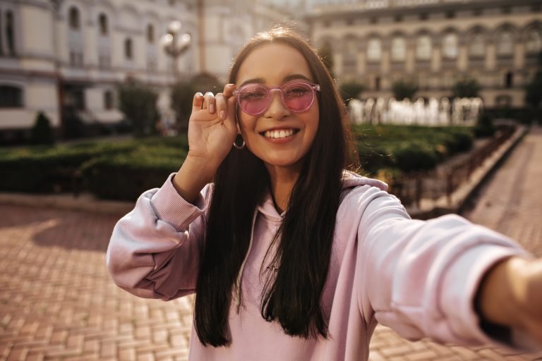 Joyful brunette woman in pink hoodie and trendy sunglasses smiles sincerely and takes selfie in goo